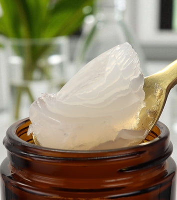 Close-up of a spoon with white cream being taken from an amber glass jar, with a blurred indoor background. Ozonated Olive Oil 